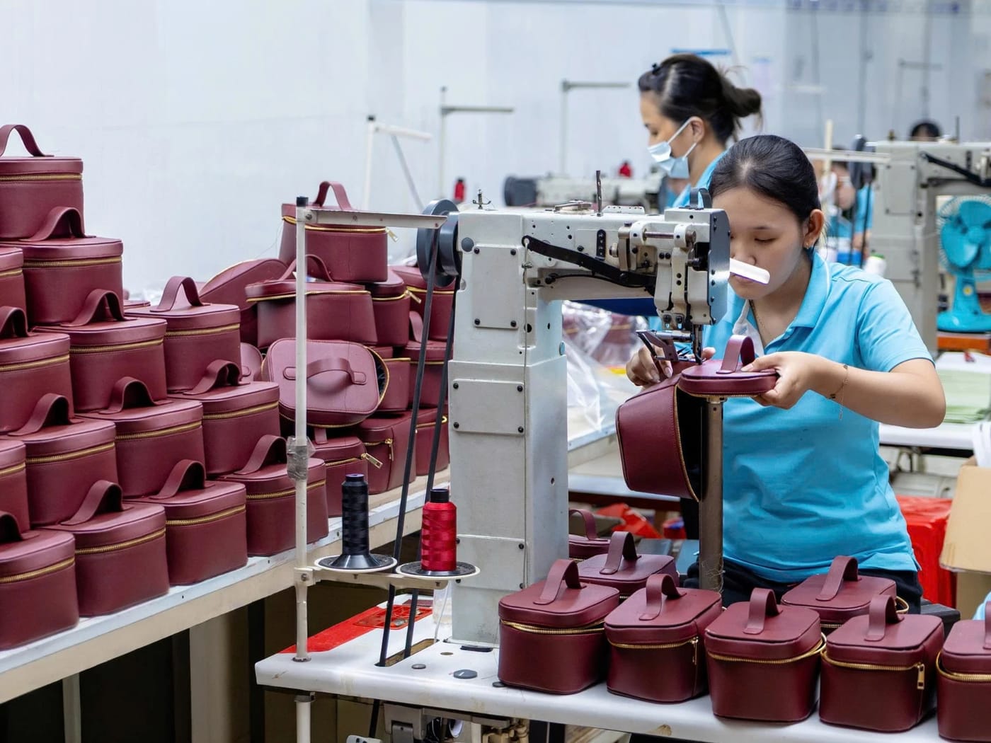 ホアンニャット工房で、職人が工業用ミシンでワインレッドのコスメポーチを縫製している様子。横には完成品が積み上げられている